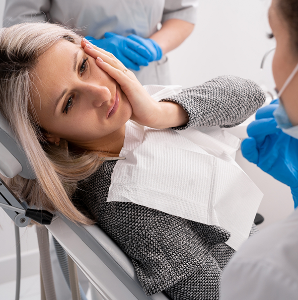 Woman in dental chair wincing and holding her cheek in pain